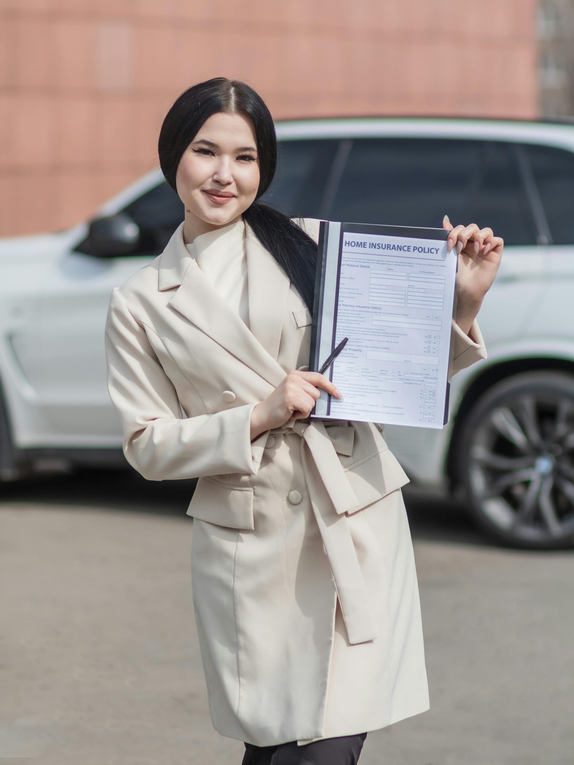 Smiling woman in beige blazer holding a home insurance policy document in outdoor setting.
